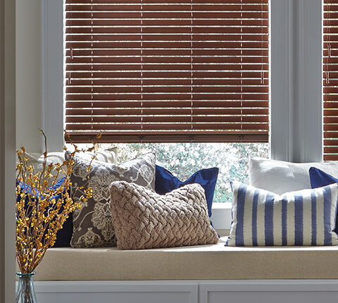 Wooden blinds partially open above a window seat with decorative pillows and a vase with branches.